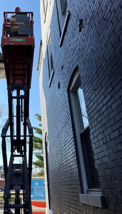 A technician stands on a scissor lift between two brick buildings, working on an air conditioning unit under a clear blue sky in Fenton.